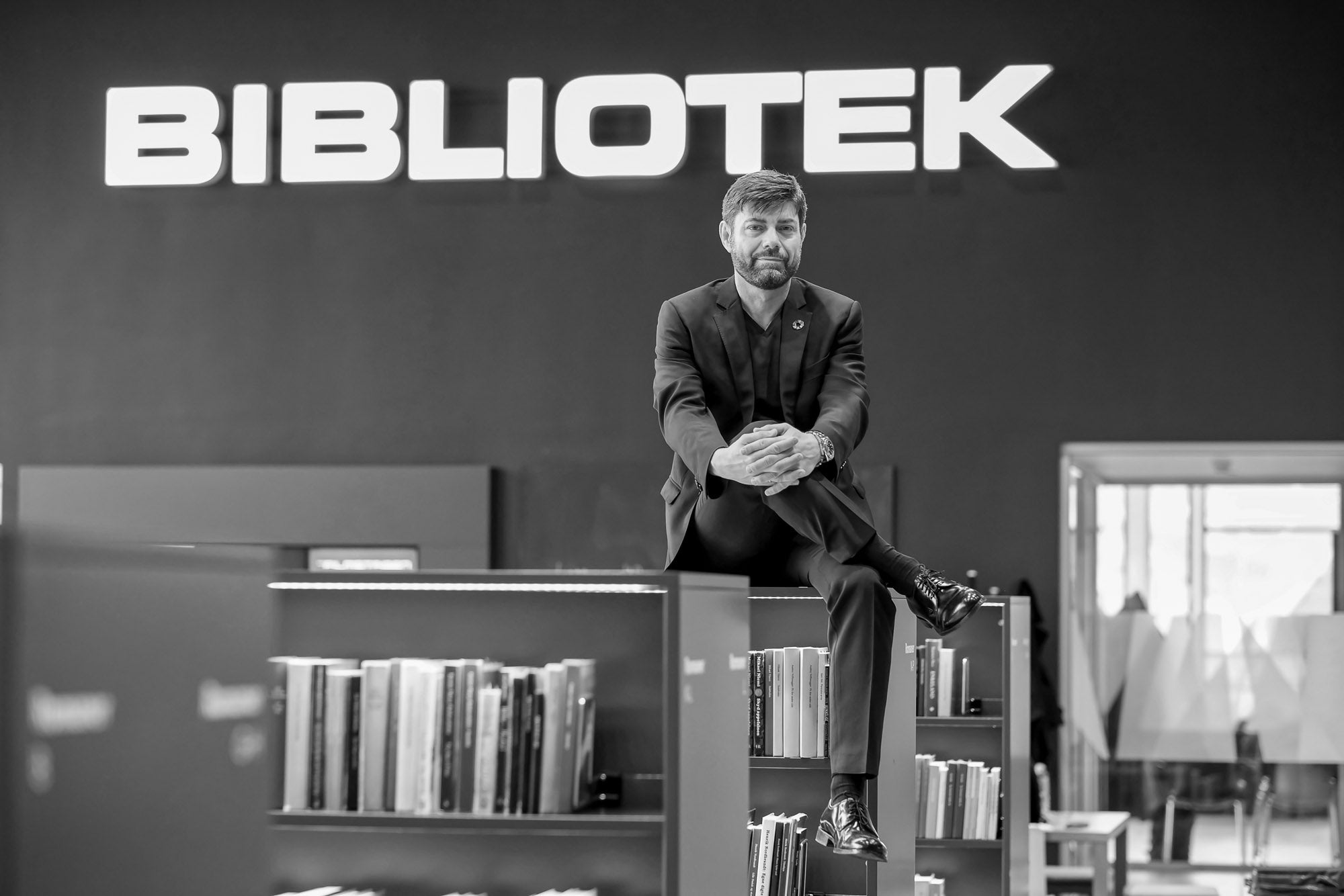 mand siddende på boghylde med biblioteks-skilt i baggrunden