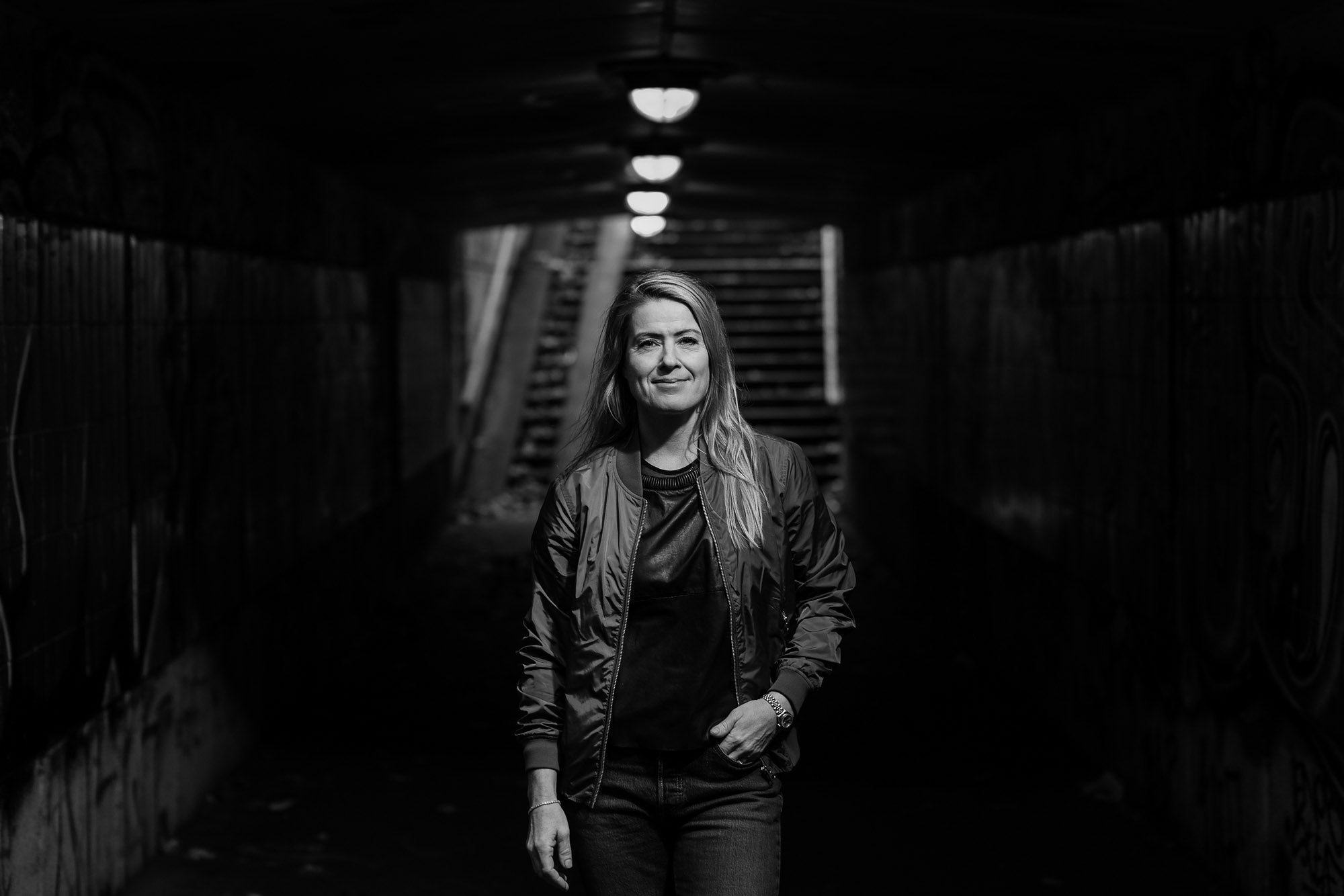 sort/hvid billede af kvinde i afslappet tøj stående i tunnel med trappe i baggrunden